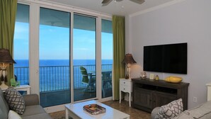 En stue med sofa, sofabord og et fjernsyn monteret på væggen. Der er en balkon med udsigt over havet.