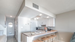 A modern kitchen with white cabinets, a central island with stools, and a dining area with a light-colored sofa. - Sea Colony 21B | Oceanfront, Private Beach Walkway (Carolina Beach)