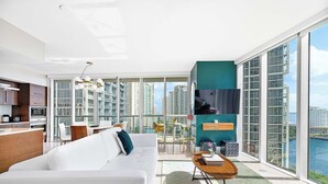Un appartement moderne comprenant un salon, un coin repas et une cuisine. Il dispose de grandes fenêtres, d’un balcon et offre une vue sur les immeubles de grande hauteur.