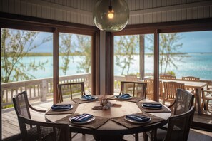 A round dining table with eight chairs, set with blue napkins and plates, overlooking a beach with clear water and trees. - The Lodge at Parrot Cay, TCI (Caicos Islands)