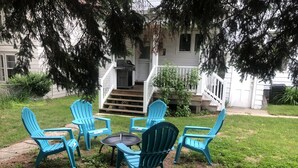 Un patio con cuatro sillas Adirondack azules y una pequeña mesa redonda, rodeado de césped, árboles y un edificio blanco al fondo.