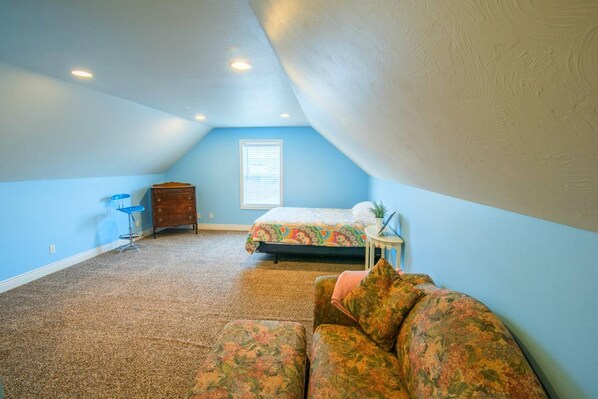 A bedroom with a sloped ceiling, a bed, a dresser, a chair, and a window.