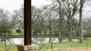 A covered porch with a stone pillar, a concrete floor, and a view of a grassy area with trees and a body of water in the background.