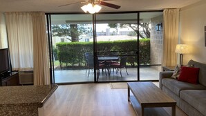 A living room with a sofa, coffee table, and a dining area with chairs and a table. There is a ceiling fan, a TV, and a lamp. - Read our Reviews! Newly Updated Condo! Perfect Getaway Steps to Beach & Dining! (Kihei)