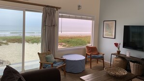 Ein Wohnzimmer mit großem Fenster mit Blick auf einen Strand, möbliert mit einer Couch, Sesseln und einem Couchtisch.