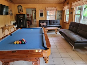 Una mesa de billar con bolas, un sofá y un soporte para tacos, todo en una habitación con paredes de madera y piso de baldosas.