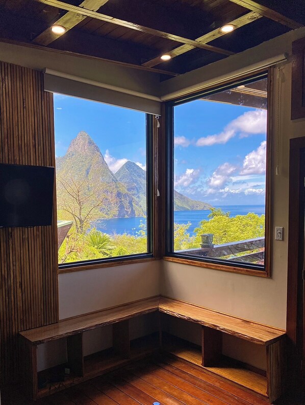 A corner room with wooden flooring, a bench, and large windows offering a view of mountains and a body of water.