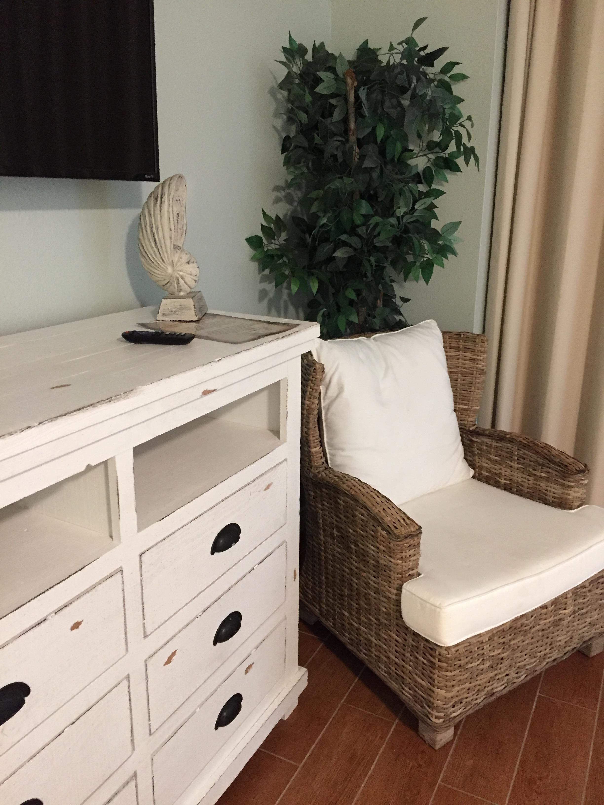 A wicker chair with a white cushion, a white dresser with drawers, a decorative shell, and a green plant.
