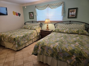 A bedroom with two beds, a nightstand, a lamp, a window with blinds, and wall decorations.