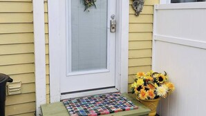 Eine Haustür mit weißem Türblatt und Glasfenster, eine kleine Veranda mit einer farbenfrohen Fußmatte und ein Blumentopf mit gelben und orangen Blumen.