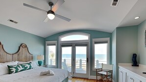 A bedroom with a bed, a ceiling fan, a view of the beach, a chair, and a desk. - A Shore Cure | Panoramic Ocean Views (Kure Beach)
