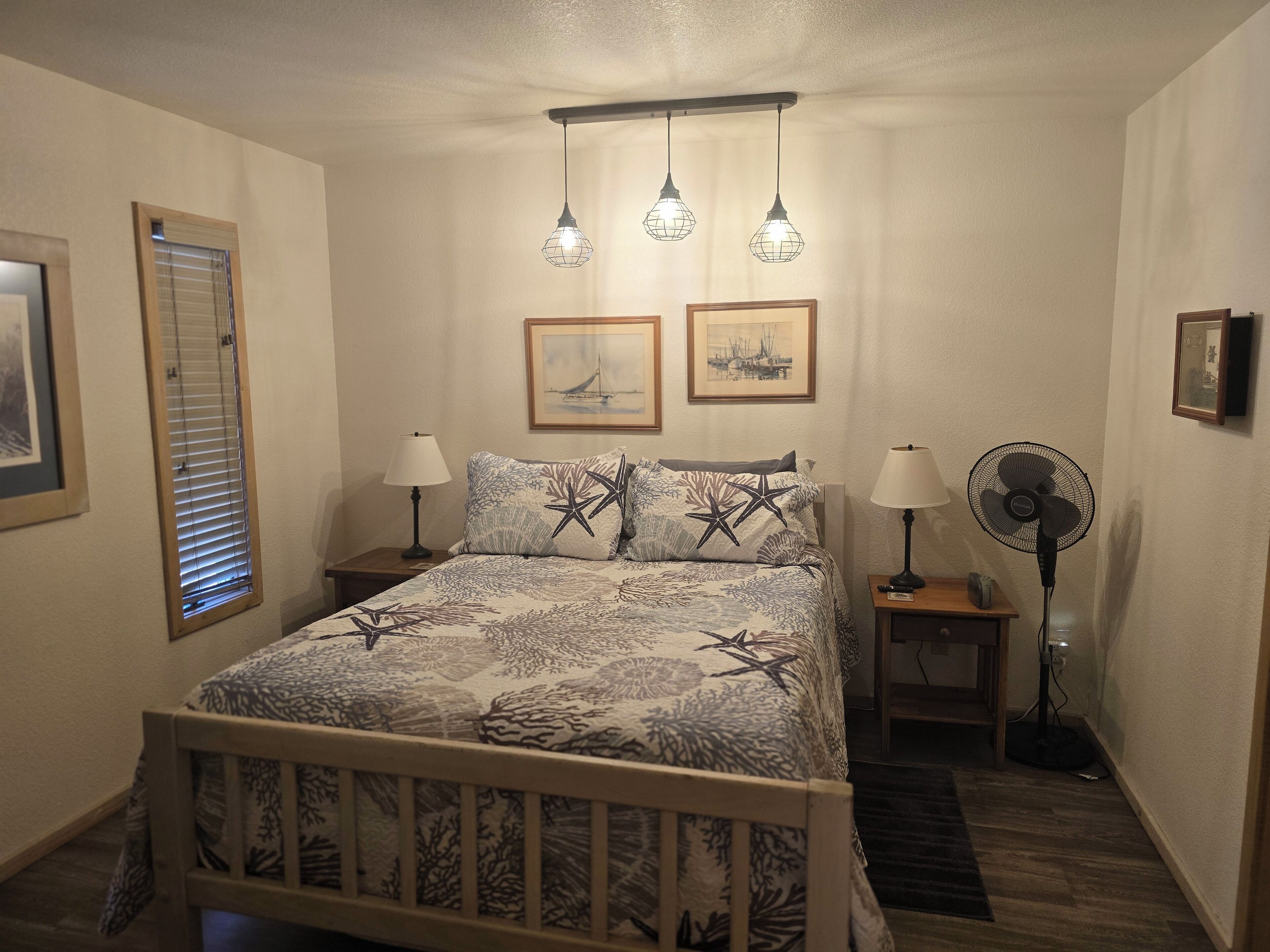 A bedroom with a bed, two bedside lamps, a standing fan, and two framed pictures on the wall.