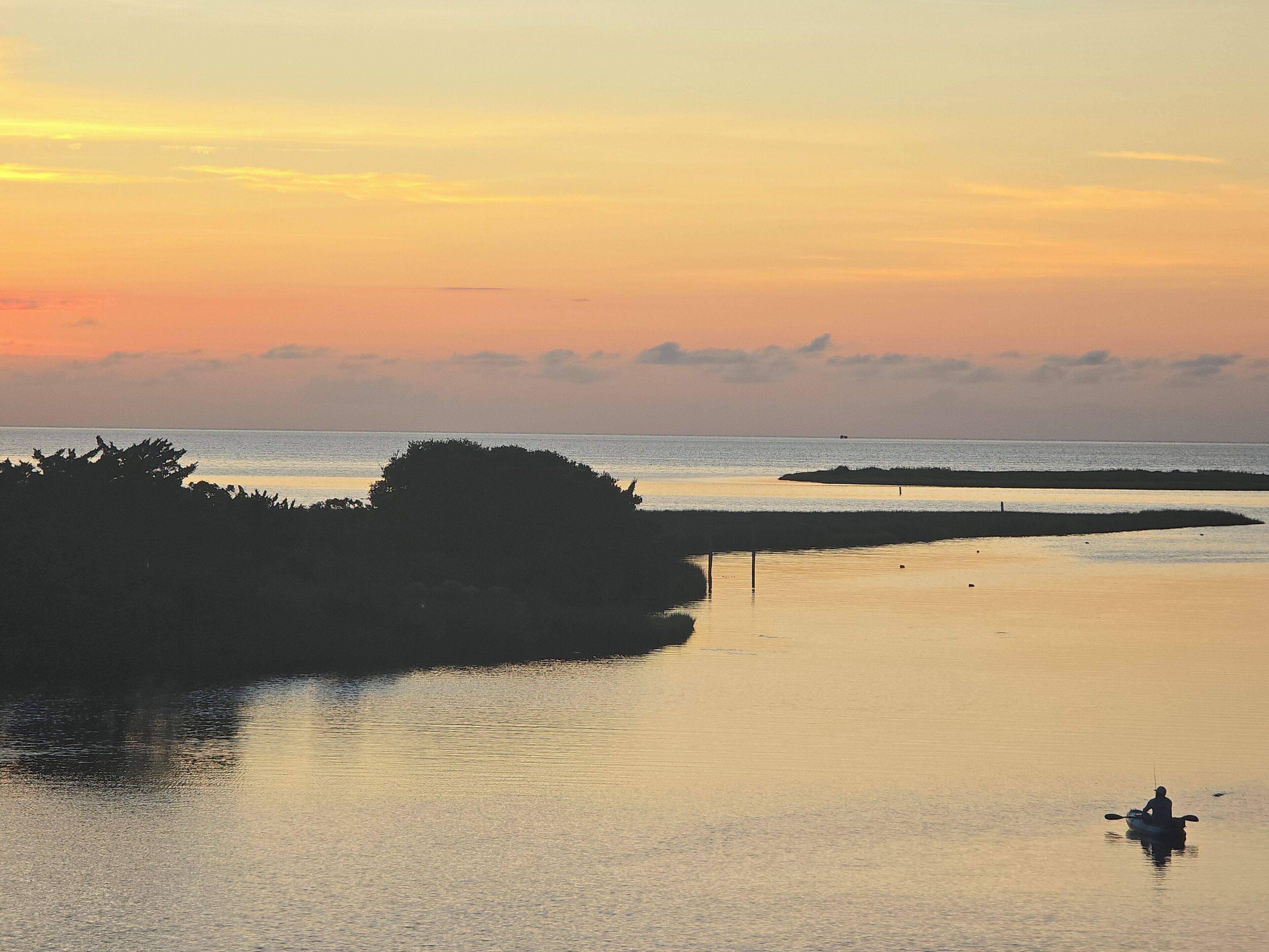 A calm body of water with a silhouette of a person in a kayak, surrounded by natural landforms and a sunset sky.