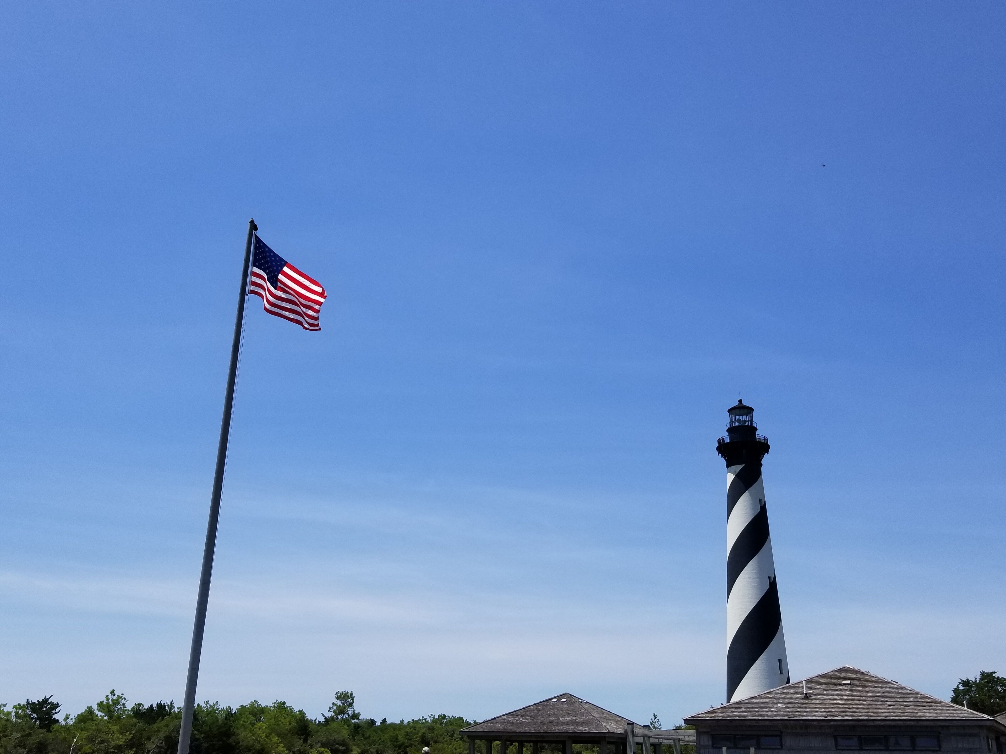 Un phare aux rayures noires et blanches, un mât portant le drapeau américain et un ciel d’un bleu limpide.