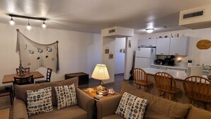 Ein Wohnzimmer mit einer Couch, Sesseln, einem Couchtisch und einem Essbereich mit Tisch und Stühlen. Es gibt eine Küchenzeile mit Schränken und Geräten.