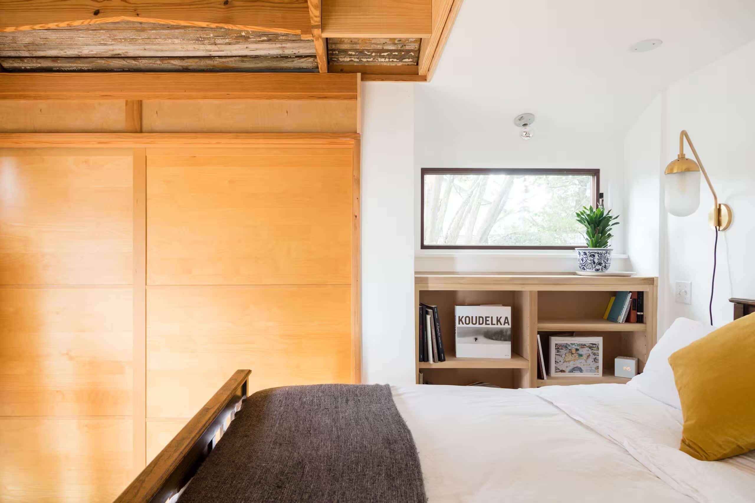 Une chambre à coucher avec un lit, un bureau agrémenté d’une plante, des livres et une fenêtre.