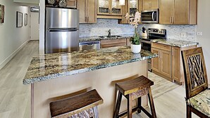 A kitchen with wooden cabinets, a stainless steel refrigerator, granite countertops, and bar stools. - Spacious Kihei Remodeled Condo New AC Waterfall Pool Steps to Kamaole Beach (Kihei)