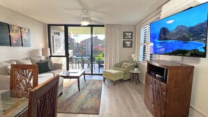 A spacious living room with a sofa, a coffee table, a TV, and a balcony with a view of buildings and greenery. - Spacious Kihei Remodeled Condo New AC Waterfall Pool Steps to Kamaole Beach (Kihei)