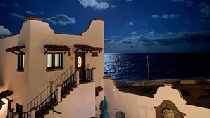 A coastal building with a staircase leading to a balcony, a clock, and a view of the sea.