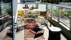 A balcony with a glass railing, a table with chairs, a patterned rug, and a small stool. - WEST OCEAN TOWERS Long Bch Condo 2Bed/2BA/Sunny/Balcony/ Pool/Gym/BEST BUILDING! (Long Beach)
