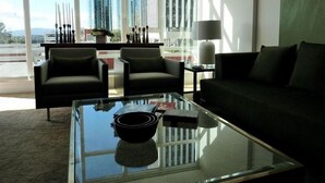 A modern living room with a glass coffee table, two armchairs, a sofa, and a view of tall buildings through large windows. - WEST OCEAN TOWERS Long Bch Condo 2Bed/2BA/Sunny/Balcony/ Pool/Gym/BEST BUILDING! (Long Beach)