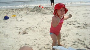 A beach scene with a child in a red swimsuit and a red hat, a person in the background, and a sandcastle. - Peaceful Villa with Ocean and Hunting Island View (Harbor Island)