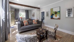 Un salon moderne avec un canapé gris, un pouf à motifs et une petite table. Un tableau est accroché au mur, et il y a une fenêtre avec des cadres blancs.