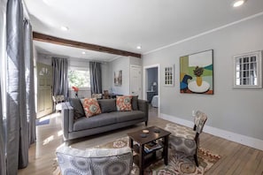 Ein modernes Wohnzimmer mit einem grauen Sofa, einem gemusterten Hocker und einem kleinen Tisch. An der Wand hängt ein Gemälde und ein Fenster mit weißen Rahmen.