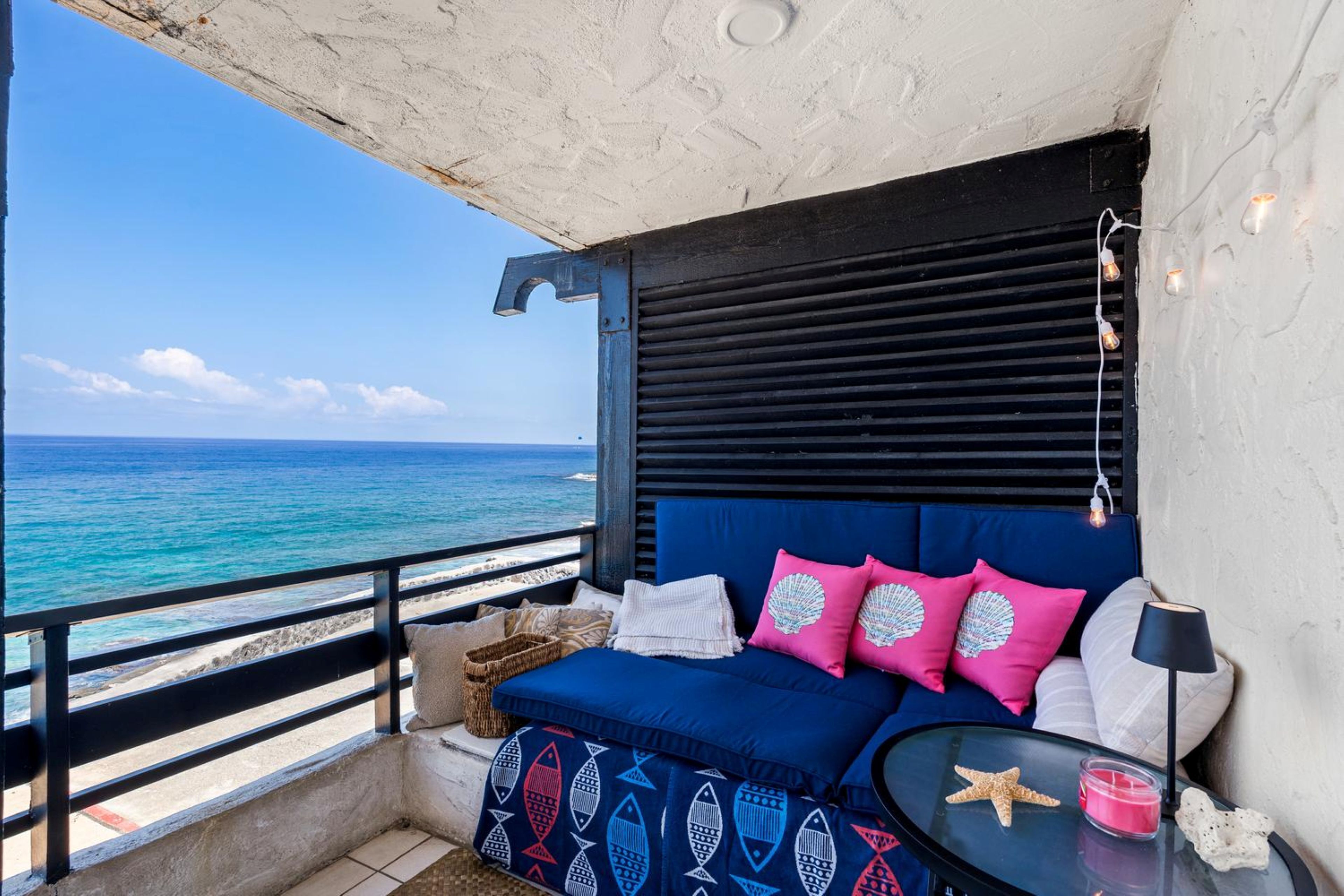 A balcony with a view of the ocean, a blue sofa with pillows, a starfish decoration, and a small table with a candle.