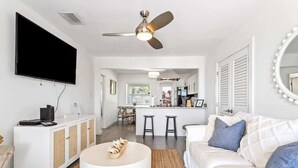 Een moderne woonkamer met een flatscreen televisie, een plafondventilator, een ronde salontafel, een witte bank en een keuken in de achtergrond.