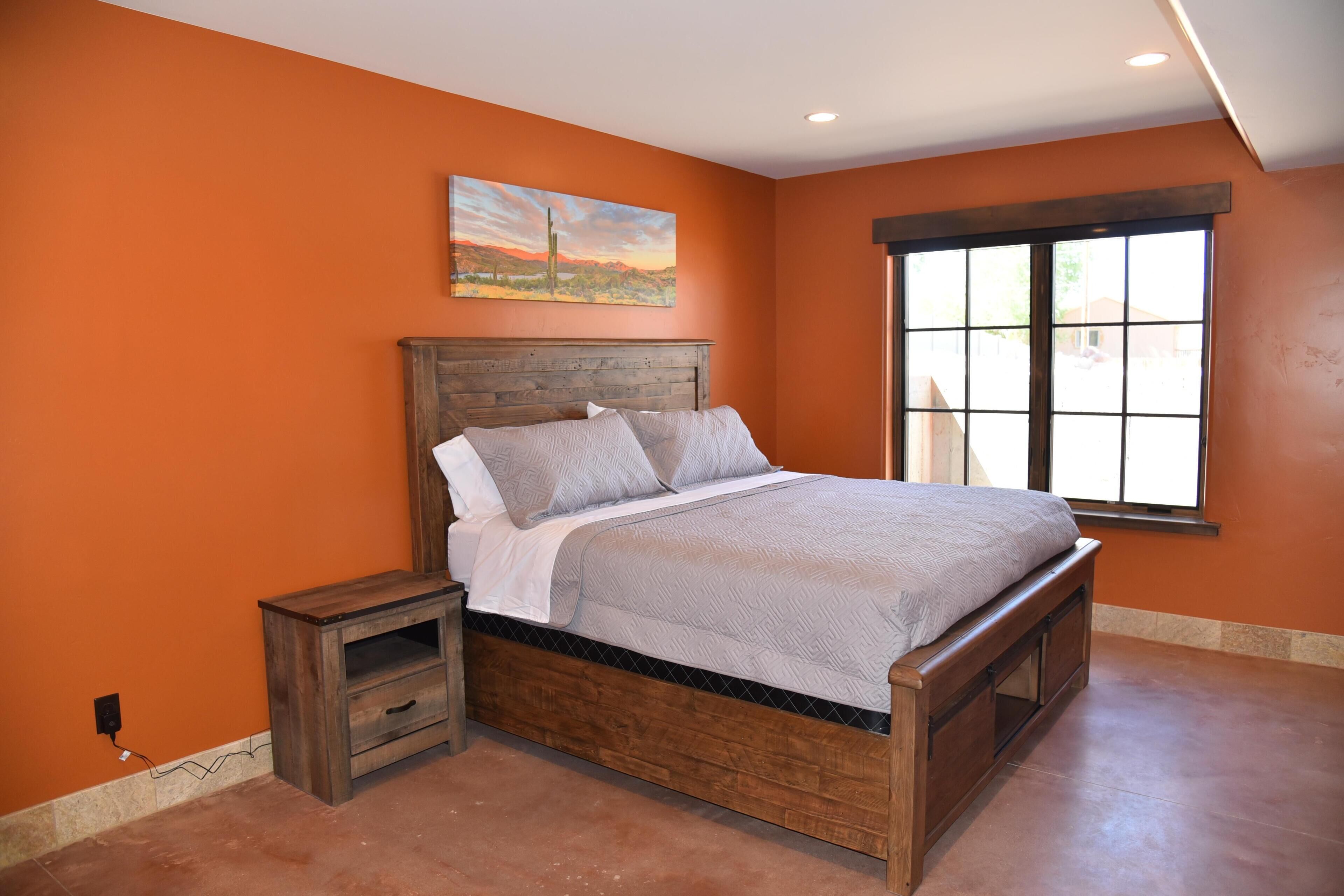 Une chambre avec un mur orange, un cadre de lit en bois, une table de chevet et une fenêtre donnant sur un beau paysage.