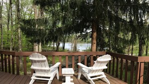 Een houten terras met twee witte Adirondack-stoelen en een kleine tafel, met uitzicht op een bos en een waterlichaam.