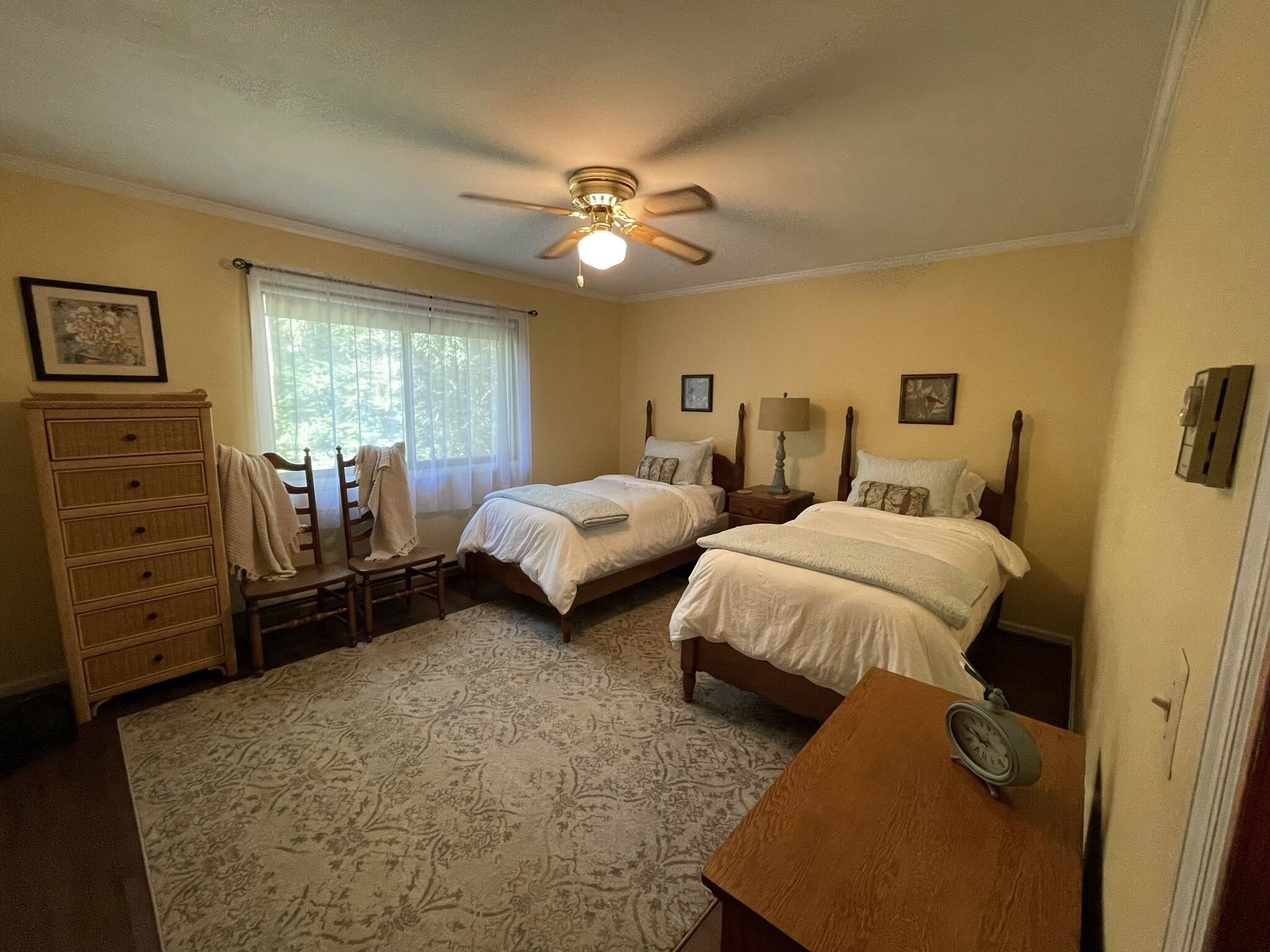 A bedroom with two beds, a dresser, a chair, a desk, a ceiling fan, and a window with curtains.