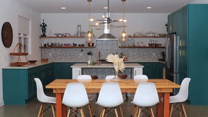 Eine moderne Küche mit türkisfarbenen Schränken, einem Holztisch, weißen Stühlen und einer zentralen Kochinsel mit einer Vase getrockneter Blumen.