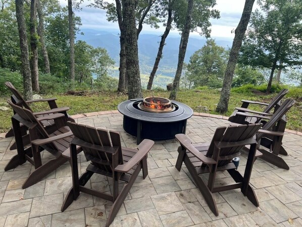 Ein runder Sitzbereich im Freien mit Feuerstelle, umgeben von vier Adirondack-Stühlen, auf einer Steinterrasse mit schöner Aussicht auf Bäume und Berge im Hintergrund.