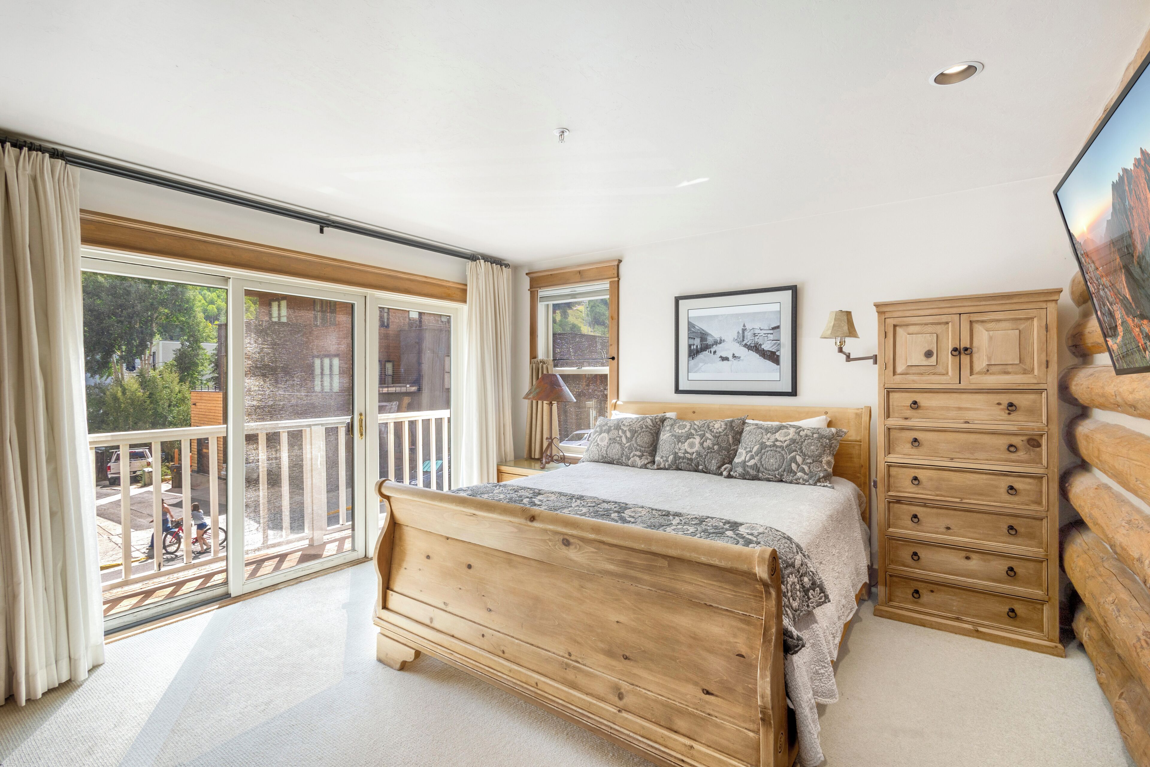 A bedroom with a large bed, a wooden dresser, a framed picture on the wall, and a balcony with a view of the outside.