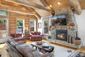 A log cabin interior with a stone fireplace, leather sofas, and a large window overlooking the outdoors. - Diamondtooth 6, Downtown Telluride Luxury Condo with Private Hot Tub, Garage (Telluride)