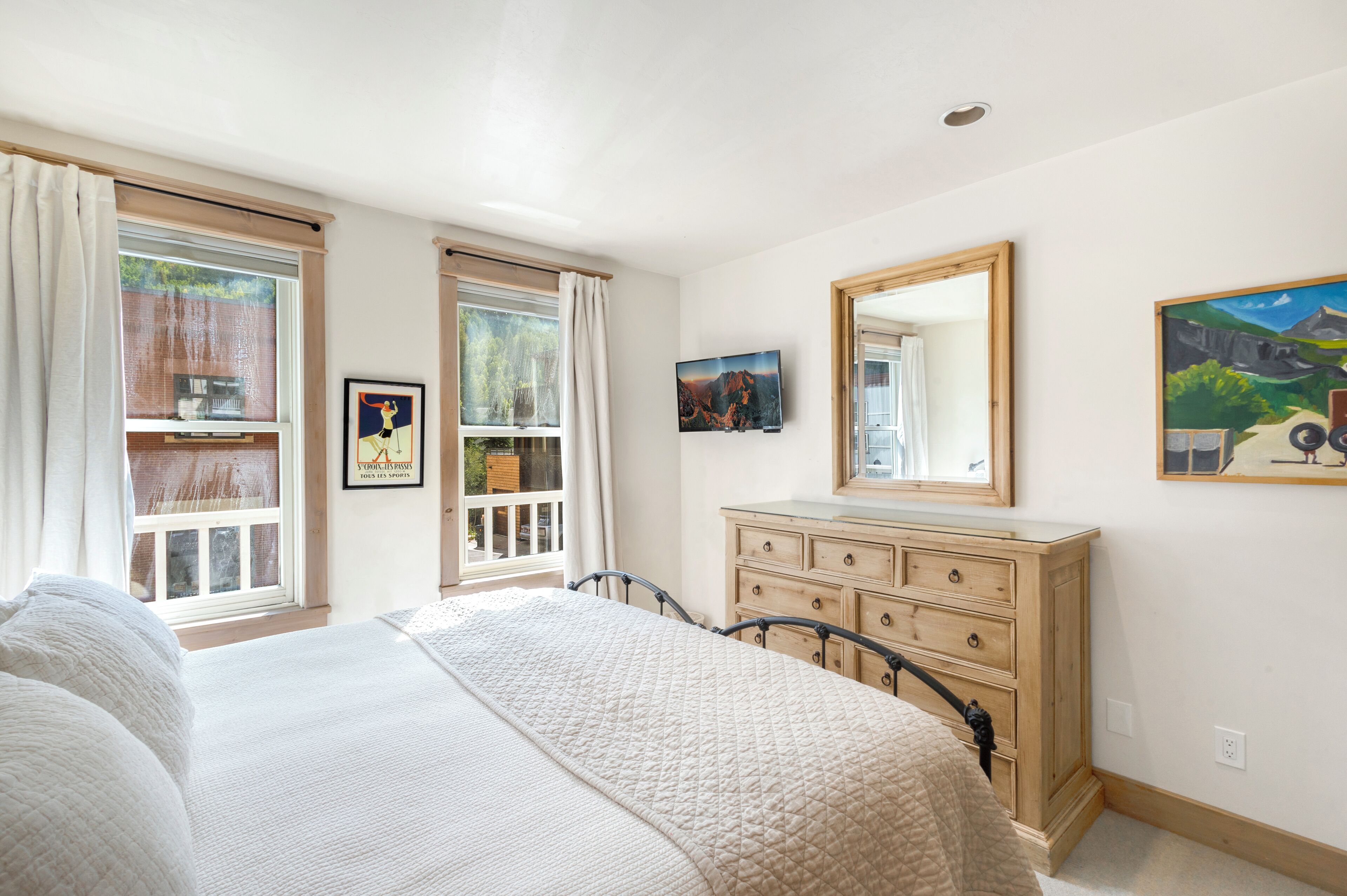 A bedroom with a bed, dresser, and a large mirror. There are windows with curtains, a painting on the wall, and a framed picture above the dresser.
