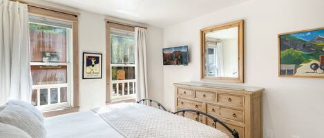 A bedroom with a bed, dresser, and a large mirror. There are windows with curtains, a painting on the wall, and a framed picture above the dresser.