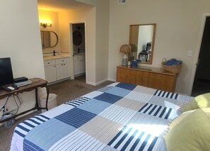 Un dormitorio con una cama, un aparador, un espejo, un escritorio con una computadora y un baño a simple vista al fondo.