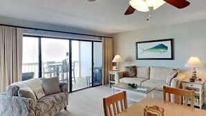 A living room with a sofa, a coffee table, a dining table, and a ceiling fan. There is a large window with a view of the outside. - Sea Colony 12A | Oceanfront + Private Beach Walkway (Carolina Beach)