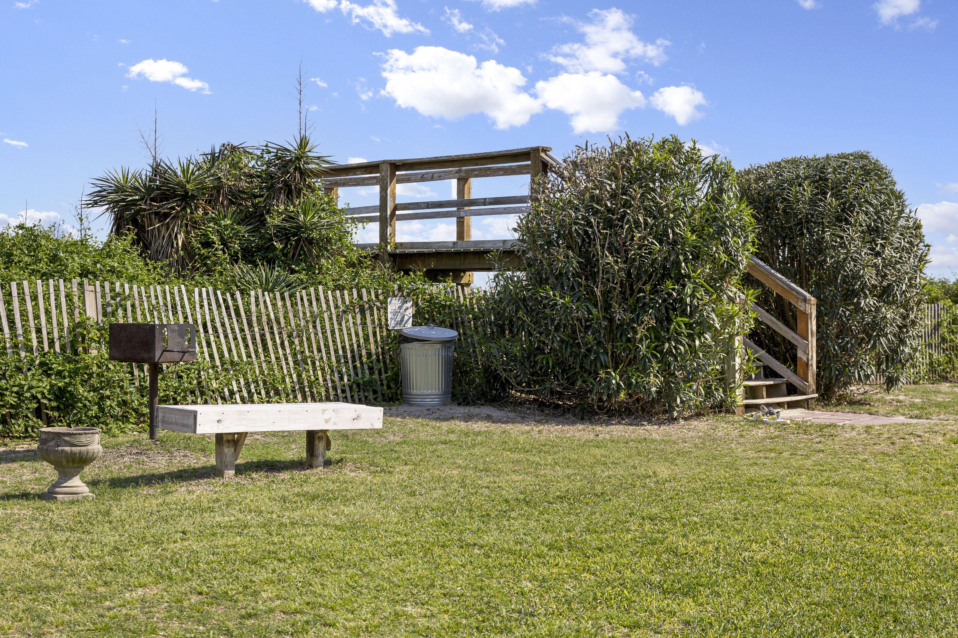 Eine Holzterrasse mit einer Bank und einem Mülleimer, umgeben von üppigem Grün und blauem Himmel.