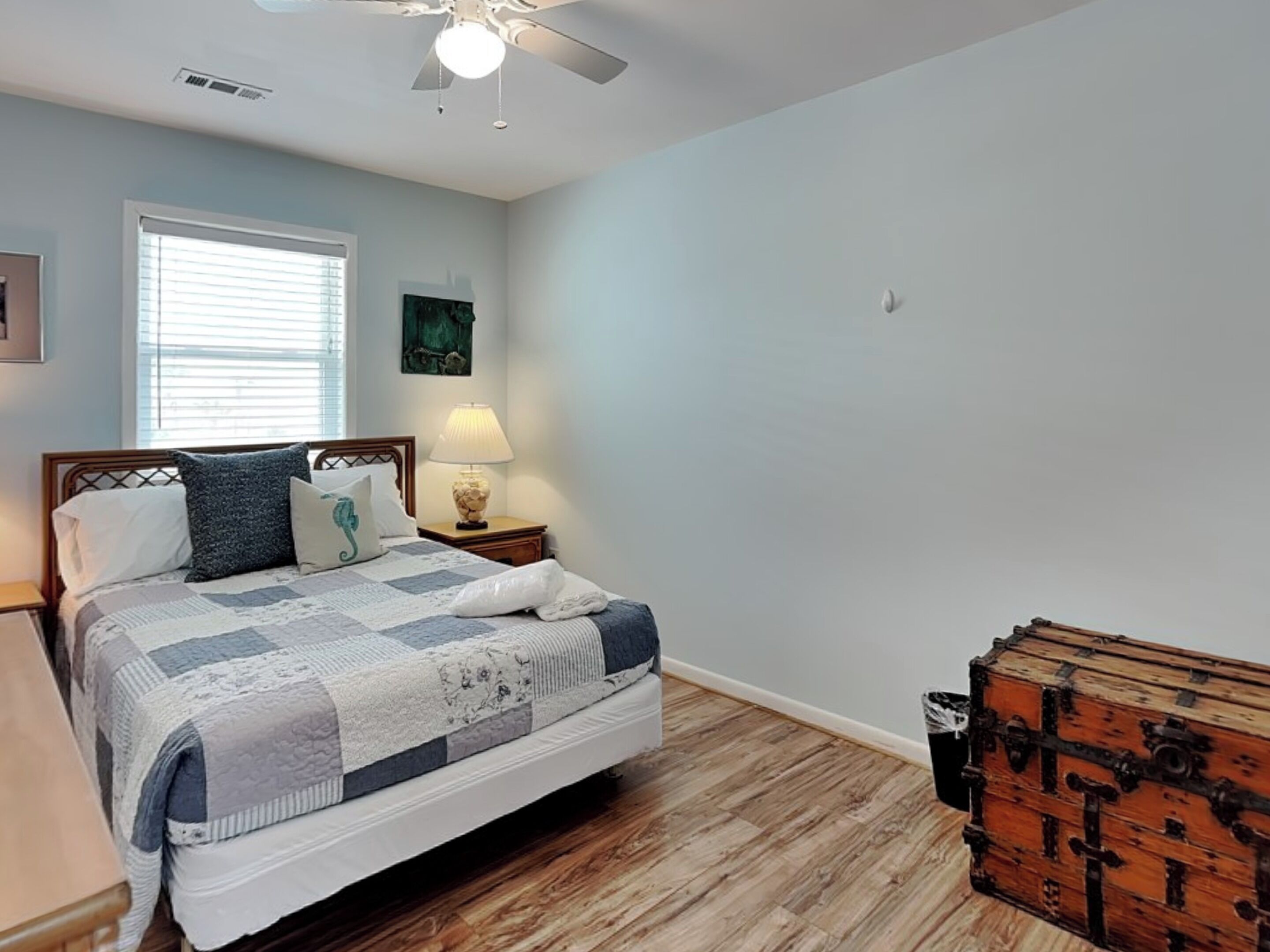 A bedroom with a bed, a nightstand, a lamp, a ceiling fan, a window with blinds, and a wooden chest.