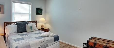 A bedroom with a bed, a nightstand, a lamp, a ceiling fan, a window with blinds, and a wooden chest.