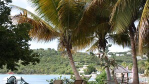 A beachside seating area with chairs and tables, palm trees, and a view of the sea and hills in the background. - String of Pearls Cozy Romantic Beach Front 1bedroom- Last Minute Discount! Ask! (Chocolate Hole)