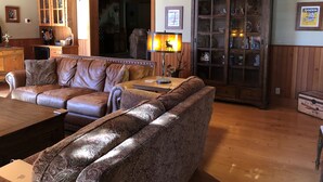 Ein gemütliches Wohnzimmer mit Ledersofas, einem Holztisch und einer Lampe. Es gibt einen Schrank mit Glastüren sowie ein Bücherregal voller Bücher und Dekoration.