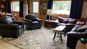 Una habitación acogedora de estilo cabaña con interior de madera, alfombra estampada, mobiliario de cuero y vistas al exterior a través de un ventanal.
