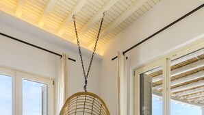 Une pièce avec un fauteuil suspendu, deux poufs en osier, et une vue sur la mer par une porte vitrée.
