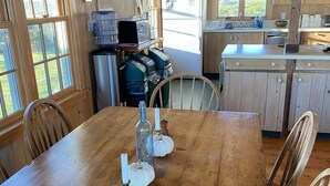 Une table de salle à manger en bois, des chaises, un réfrigérateur, un évier et une fenêtre donnant sur un paysage verdoyant.