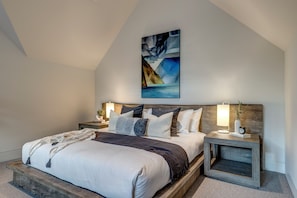 A bedroom with a sloped ceiling, a bed with a wooden headboard, two bedside tables with lamps, and a painting above the bed. - This is the largest ski in ski out rental townhome in Whistler. And it's swanky. (Whistler)